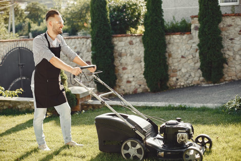 A man with a neatly groomed beard, wearing a black apron over a checkered shirt and light-colored pants, is pushing a lawn mower across a well-maintained garden lawn. The lawn mower has a black engine